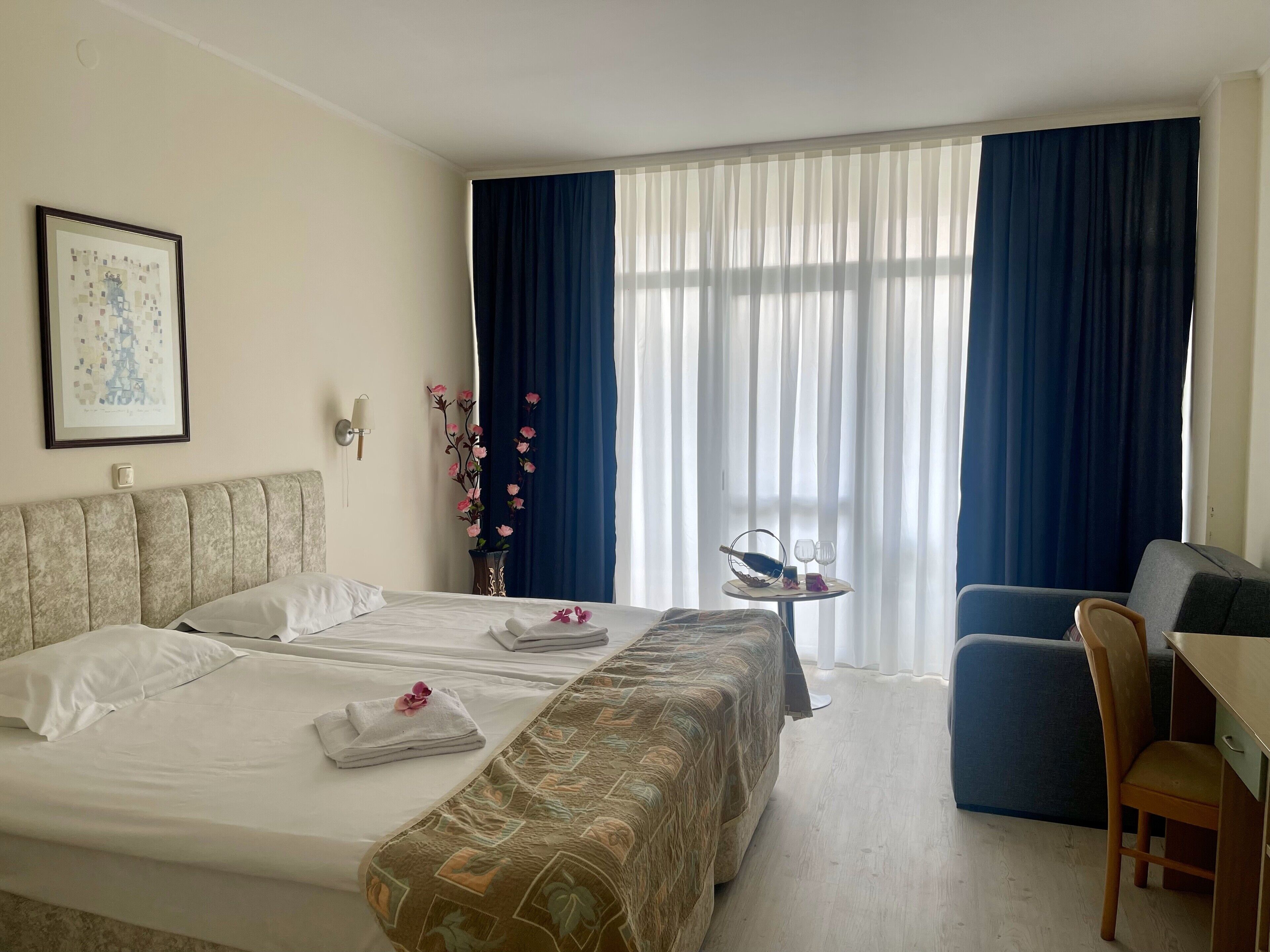 A hotel room with a bed, a desk, a chair, a vase with flowers, and a framed picture on the wall.