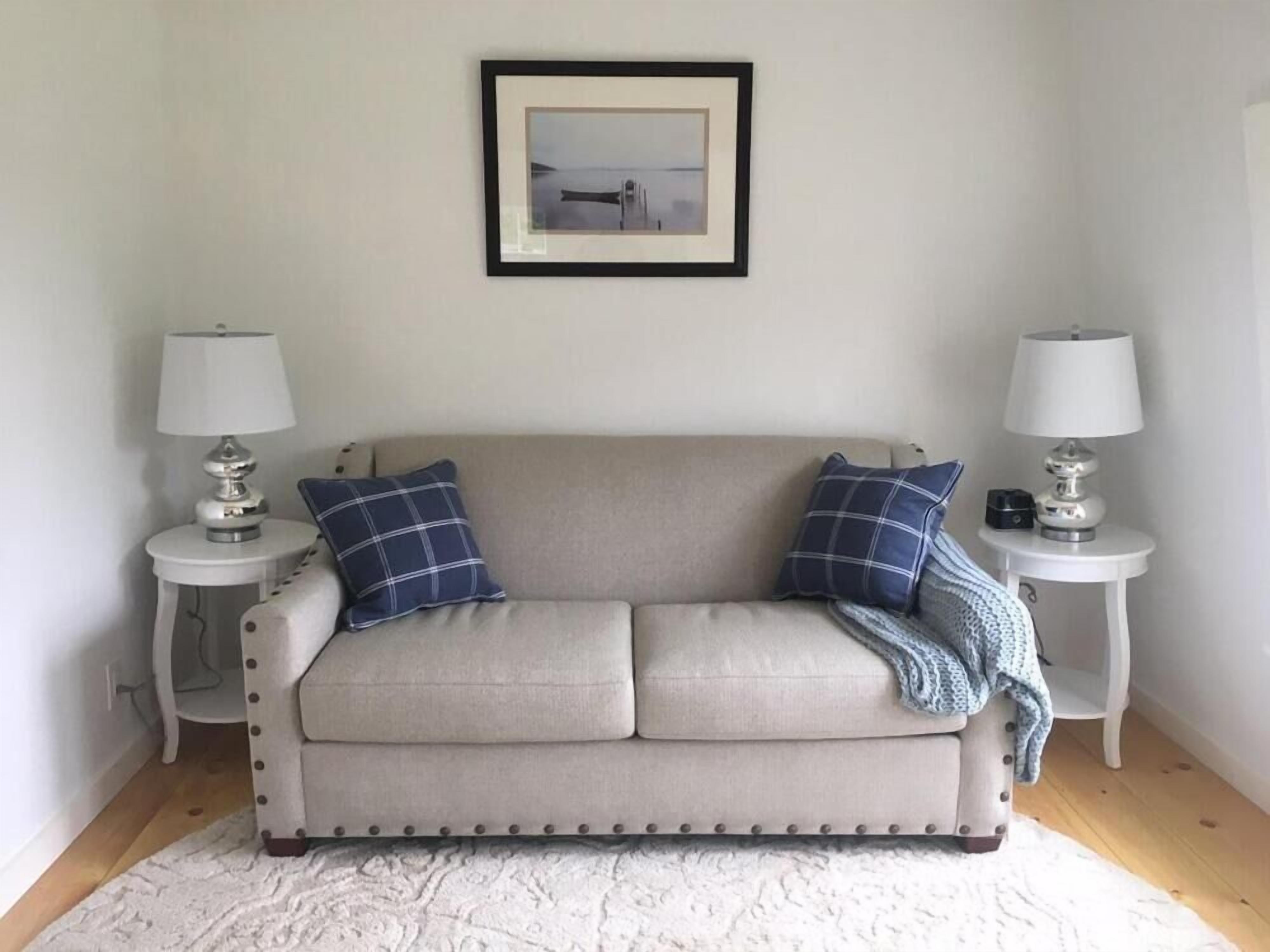 Un sofá de tonos neutros con dos almohadas a juego, flanqueado por dos pequeñas mesas blancas con lámparas, un cuadro enmarcado encima y una alfombra blanca con textura en el suelo.