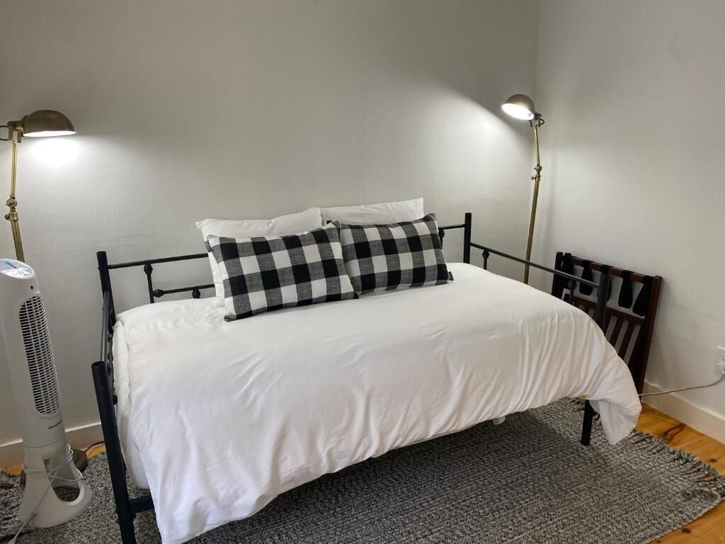 A bed with a white mattress and black and white checkered pillows, two bedside lamps, and a standing fan.