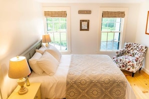Un dormitorio con cama, una mesita de noche con lámpara, dos ventanas con cortinas a cuadros, una silla y un cartel que dice 'DEER ISLE'.