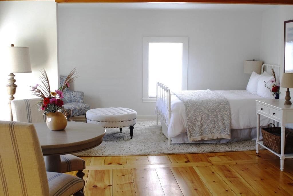 Ein Schlafzimmer mit Bett, Nachttisch, Lampe, Sessel, Tisch und Fenster.