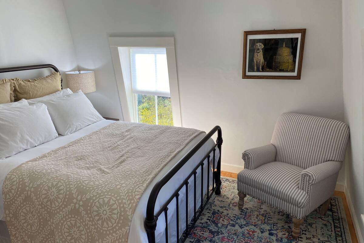 Ein Schlafzimmer mit einem Bett, einem Sessel, einem Fenster und einem gerahmten Bild an der Wand.