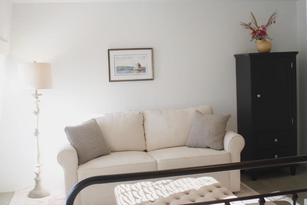 A white sofa with pillows, a framed picture on the wall, a tall floor lamp, and a black cabinet with a vase of flowers.