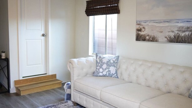 Ein beiges Sofa mit Blumenkissen, ein Fenster mit Jalousien, ein Gemälde mit Strandmotiv und eine Holztür.