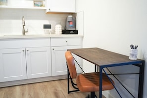 A kitchenette with a coffee maker, a small white appliance, and a white cabinet. There is a table with a chair and a cup holder with pens.