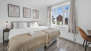 A bedroom with two beds, a window with curtains, a bedside table with a lamp, and artwork on the wall. - Modern Comfort |4BR House for 11| Central & Quite (Essex)