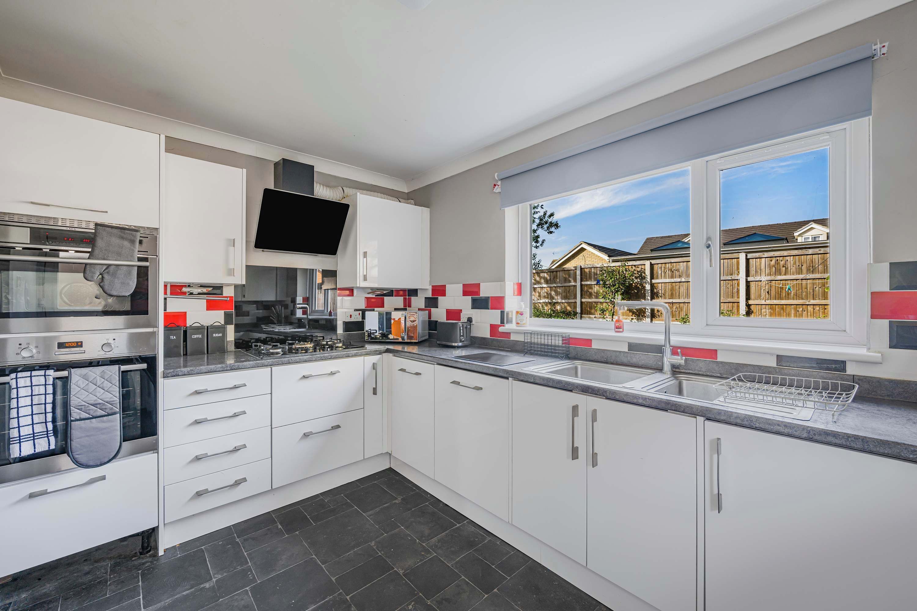 A modern kitchen with white cabinets, a built-in oven, a large window with a view of houses, and a tiled backsplash.