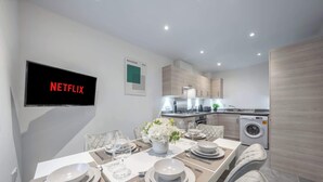 Ein moderner Essbereich mit einem Tisch für vier Personen, einem Fernseher, der 'NETFLIX' zeigt, und einer Küche im Hintergrund.