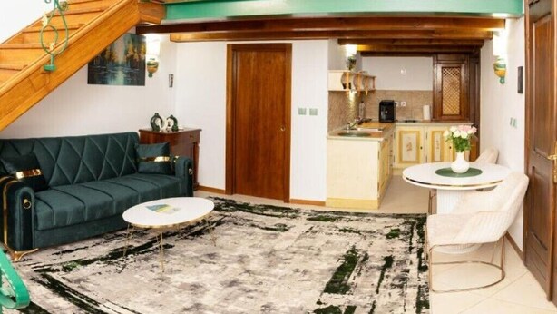 A living room with a sofa, coffee table, and a rug. There is a staircase with green railings, wooden cabinets, and a kitchen area visible. - Apartament 1 (Timișoara)