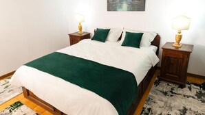 A neatly made bed with a green and white bedspread, two wooden nightstands with lamps, and a painting of trees on the wall. - Apartament 1 (Timișoara)