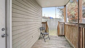Ein überdachter Balkon mit Holzterrasse, einem Metallstuhl und Blick auf einen umzäunten Garten.