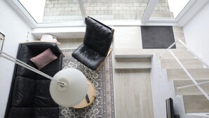 Ein modernes Interieur mit einem schwarzen Ledersofa, einer weißen Stehlampe und einem schwarzen Ledersessel. Auf dem Boden liegt ein gemustertes Teppich, und im Hintergrund ist eine Treppe zu sehen.