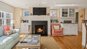 A living room with a fireplace, a television, a sofa, a coffee table, and a cabinet with glass doors. - Walk to Famous Chatham Lighthouse Beach (Chatham)