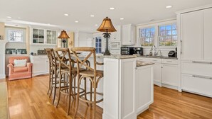 A spacious kitchen with white cabinetry, a central island with bar stools, and a dining area with a pink chair. - Walk to Famous Chatham Lighthouse Beach (Chatham)