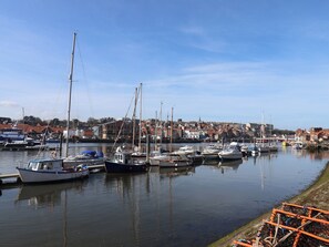 A marina with various boats docked, a calm water surface, and a town in the background. - Marina Side (Whitby)