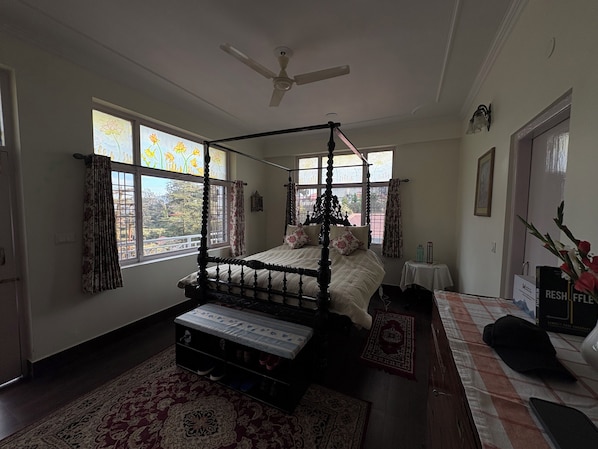 Un lit à baldaquin équipé d’un ventilateur de plafond, d’une table de chevet et d’une commode, avec vue sur l’extérieur grâce à de grandes fenêtres.