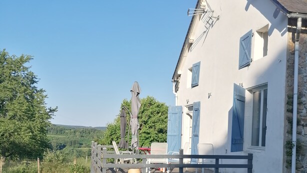 Un edificio blanco con contraventanas azules, una terraza de madera y un muro de piedra.