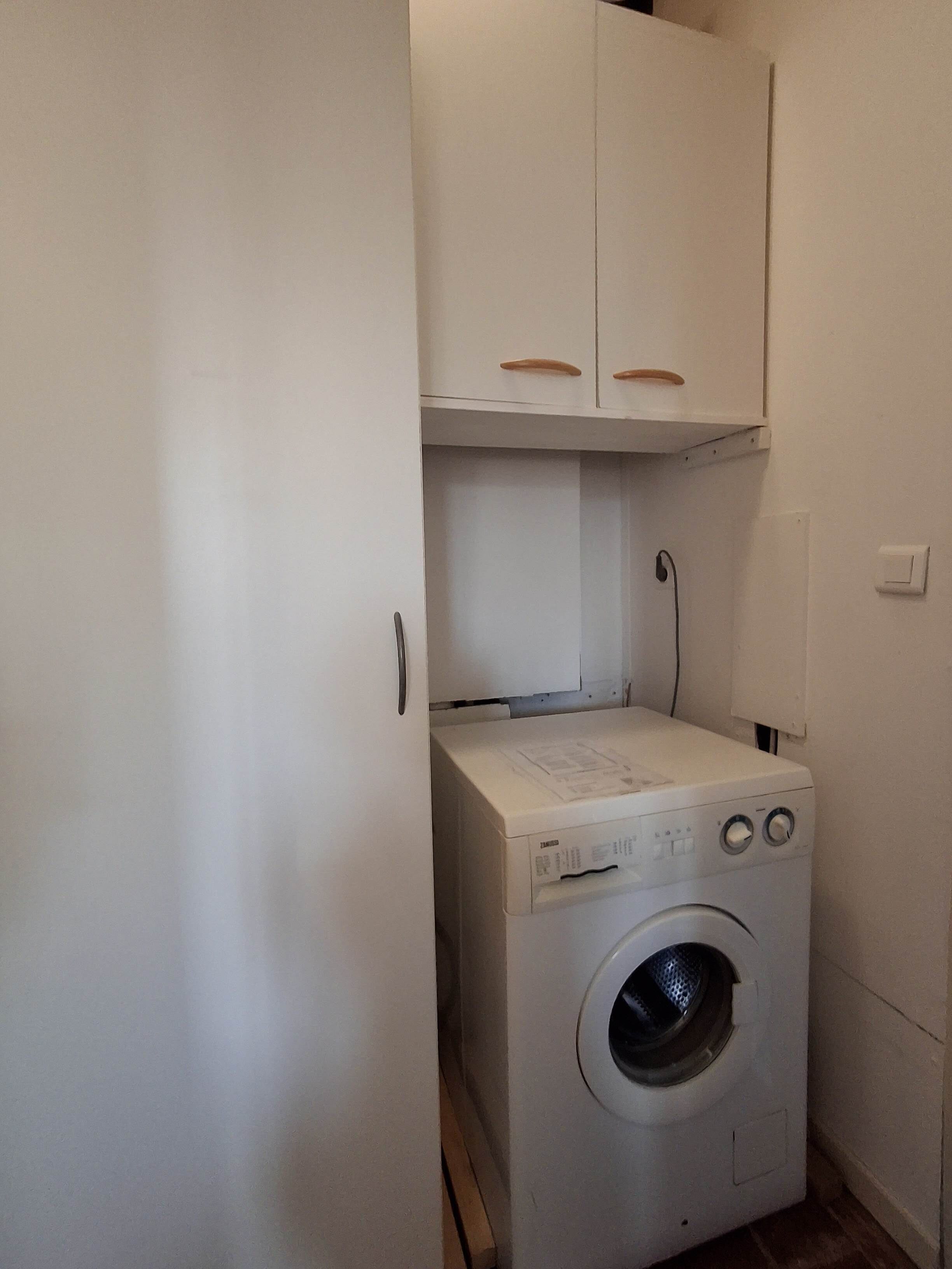 A compact laundry room with a washing machine, a cabinet above, and a power outlet on the wall.