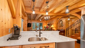 Ein Holzblockhaus-Interieur mit einer Küche, die eine Marmorarbeitsplatte, eine Kaffeemaschine, ein Waschbecken und einen Edelstahlkühlschrank zeigt.