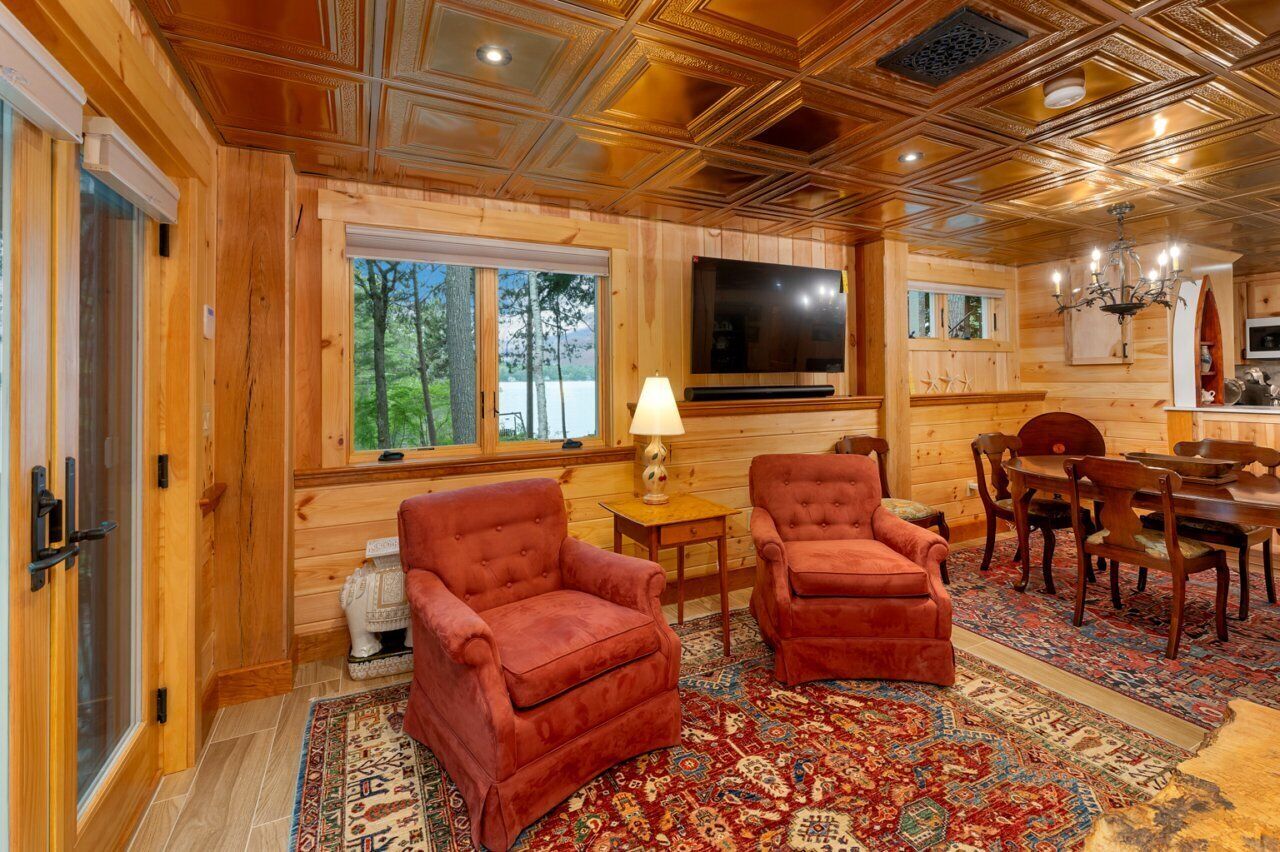 A log cabin interior with a dining area, two red armchairs, a television, and a patterned rug.
