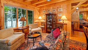 Ein Holzblockhaus im Inneren, mit einer Couch, Sesseln, einem Esstisch und einem Schrank mit gerahmten Bildern.