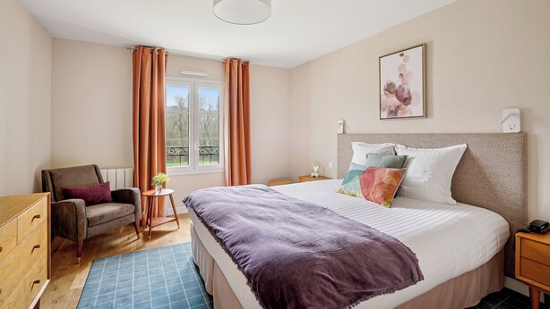 A bedroom with a bed, a chair, a dresser, a small table, and a window with curtains. - MH Hôtel & Spa (Arradon)
