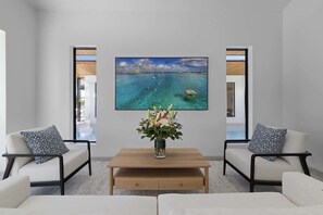 Ein modernes Wohnzimmer mit einem großen Gemälde einer Küstenlandschaft, einem Couchtisch mit Blumenvase und zwei Sofas mit gemusterten Kissen.