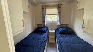 Una habitación pequeña y estrecha con dos camas individuales, una ventana con cortinas y una mesita con una lámpara.