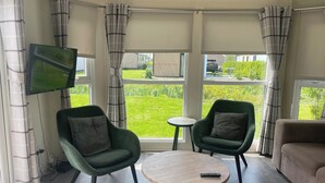 Una habitación con dos sillones verdes, una mesa redonda de madera, un televisor montado en la pared y ventanas amplias con cortinas.