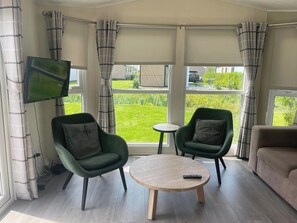 A room with two green armchairs, a round wooden table, a TV mounted on the wall, and large windows with curtains. - Getaway With Garden & Parking (West-Graftdijk)