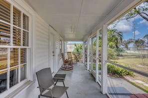 A covered porch with white siding, a bench, two rocking chairs, and a view of a garden and street.
