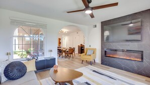 A modern living room with a fireplace, a ceiling fan, and a dining area visible through an arched doorway. - Patio & Pool Access! Mtn-view Palm Springs Escape (Palm Springs)