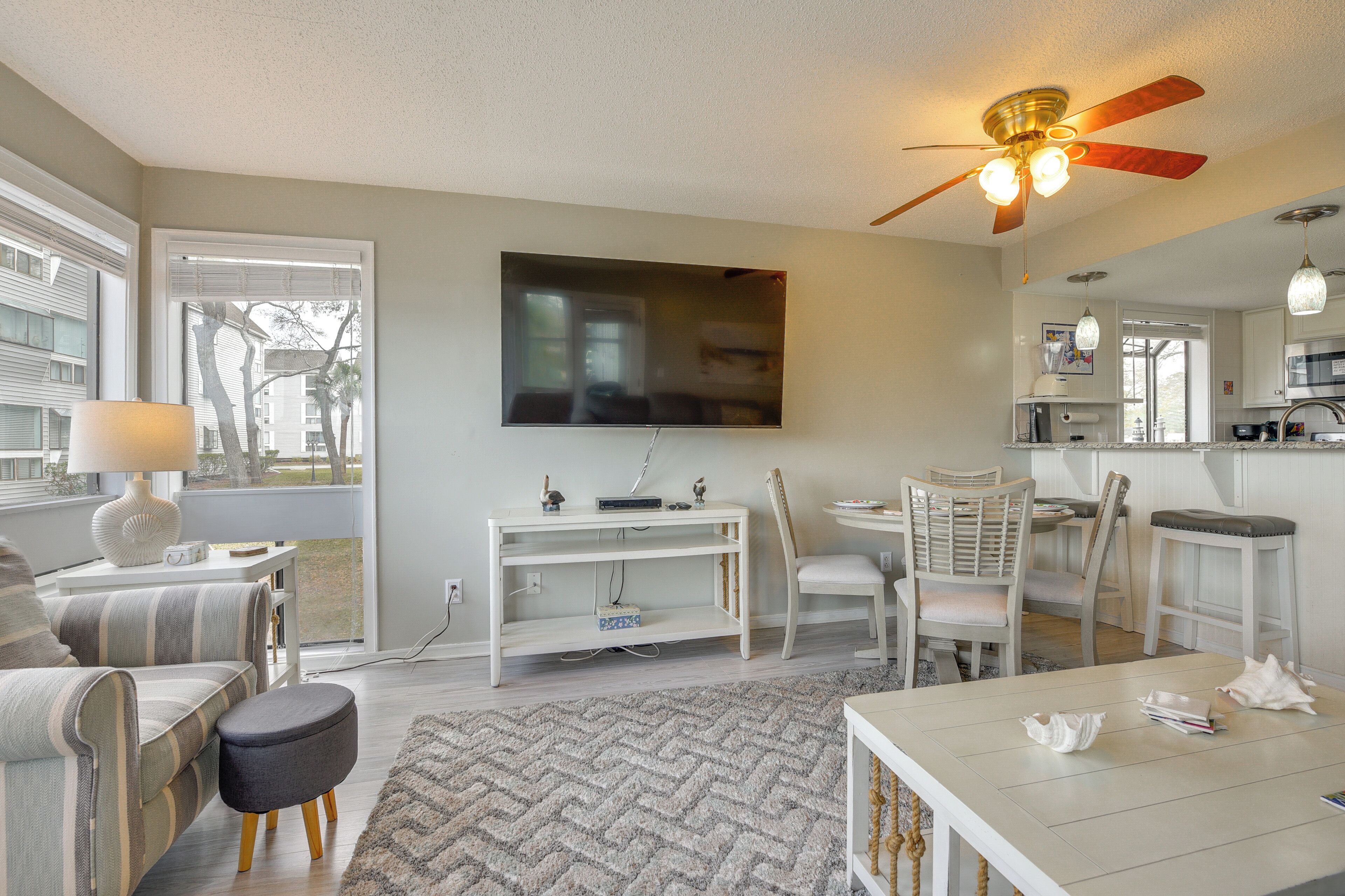 A living room with a TV, a ceiling fan, a dining area with chairs, and a kitchen area with a microwave.