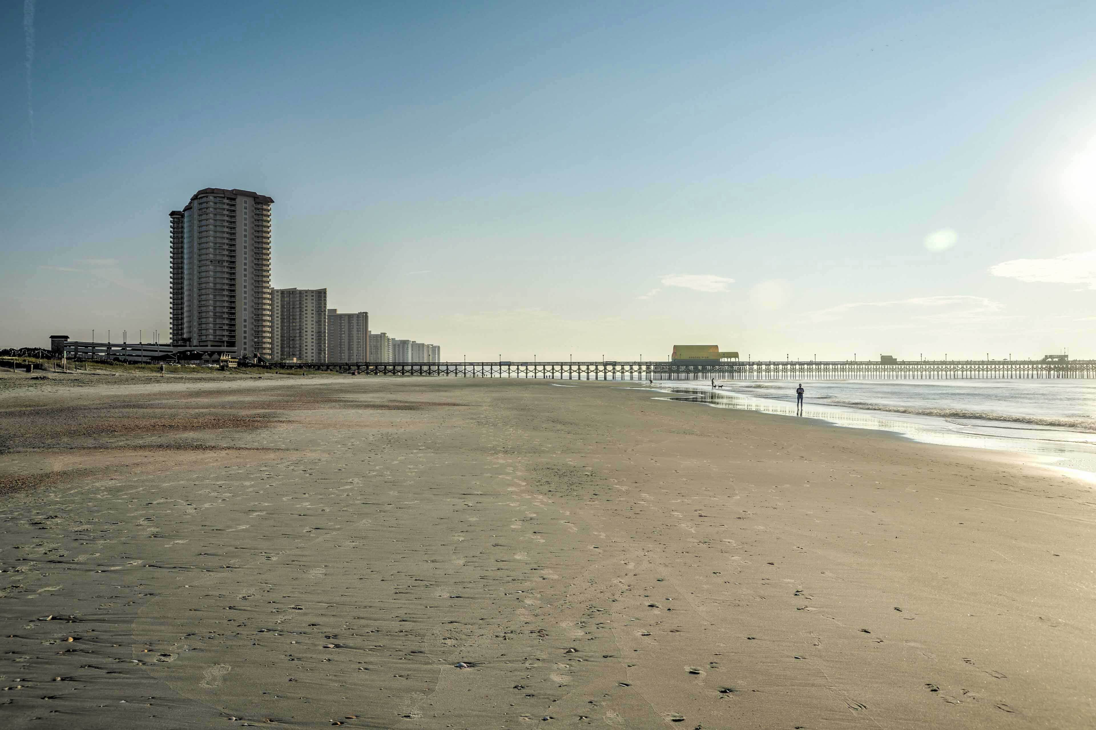 A beach with a long pier extending into the water, a tall building in the background, and a clear sky.