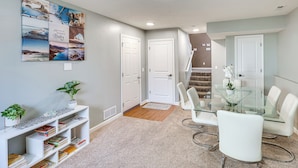A modern living room with a glass dining table, white chairs, a bookshelf with books, a wall art collage, and a staircase. - Private Yard & Patio: Spacious Retreat in Omaha! (Omaha)