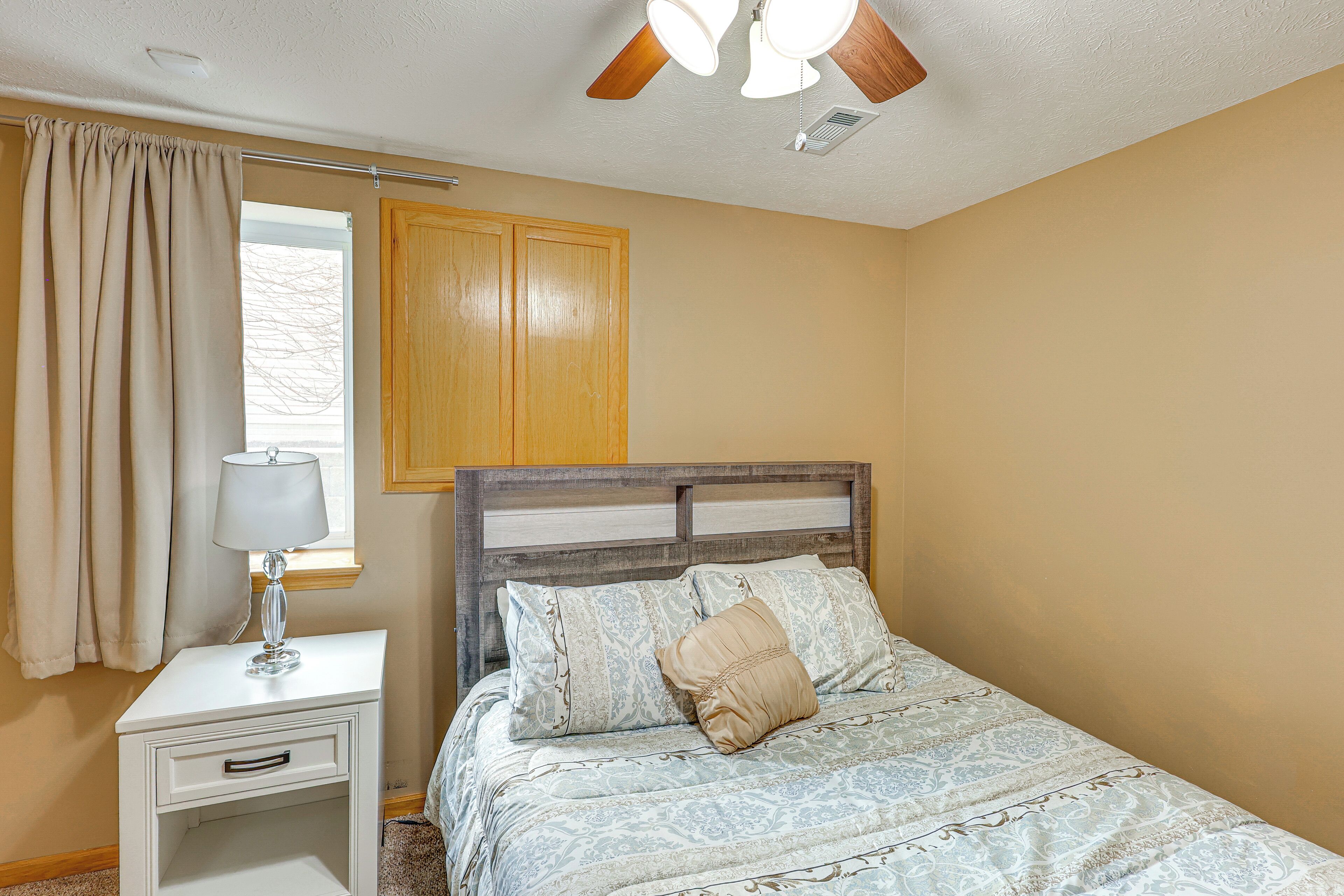 Une chambre à coucher avec un lit, une table de chevet avec une lampe, une fenêtre avec des rideaux et un ventilateur de plafond.