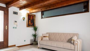 Een kamer met een beige bank, een houten plafond, een lijst met een schilderij, een plant in pot en een deur.