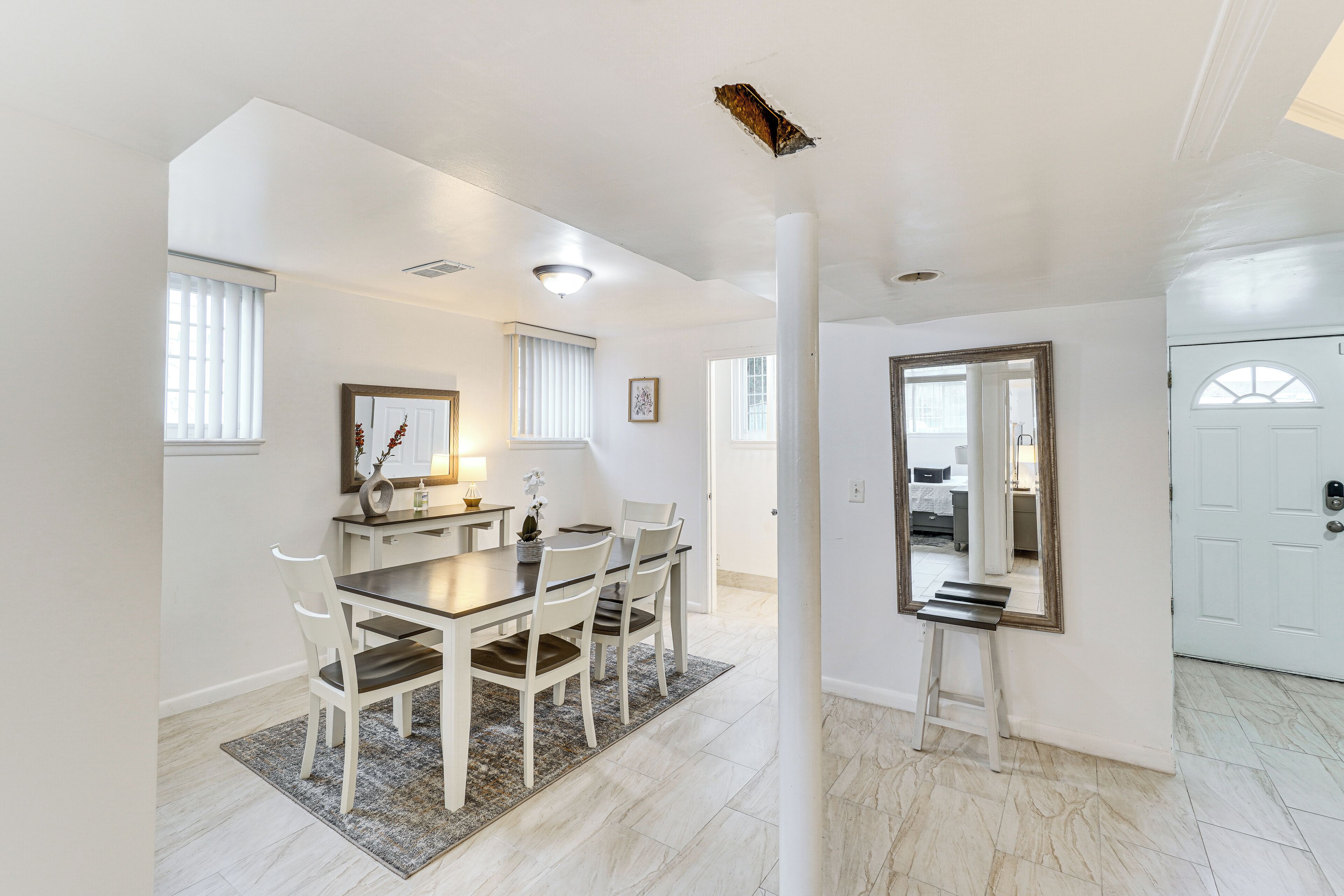 A dining area with a table, chairs, and a rug. There is a mirror on the wall and a door in the background.