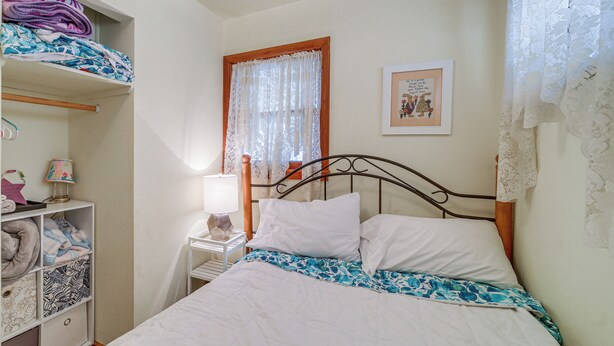 Une chambre à coucher comprenant un lit, une table de chevet, une lampe, une fenêtre avec des rideaux, un tableau accroché au mur et une étagère avec des couvertures pliées.