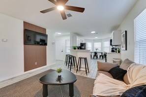 A modern living room with a ceiling fan, a TV mounted on a wooden panel, a dining area with stools, and a kitchen with a central island. - < 1 Mi to Historic Mckinney: Modern Duplex w/ Yard (McKinney)