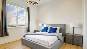 A modern bedroom with a large bed, bedside lamps, a view through a window, and a geometric light fixture. - Gateway to Route 66 Fun! Mtn-view Williams Home (Williams)