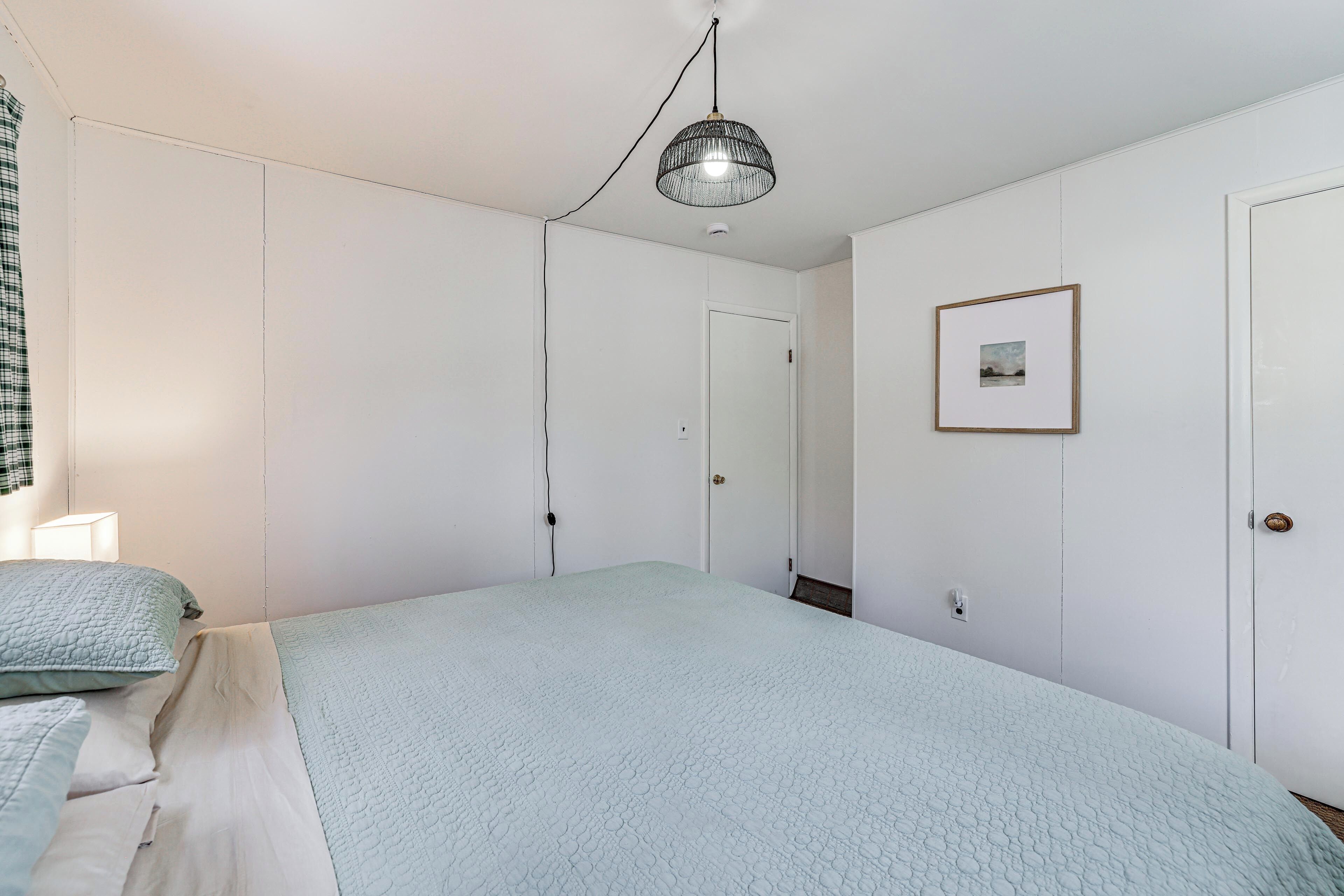 Un dormitorio con cama, lámpara de noche, lámpara colgante, puerta y un cuadro en la pared.