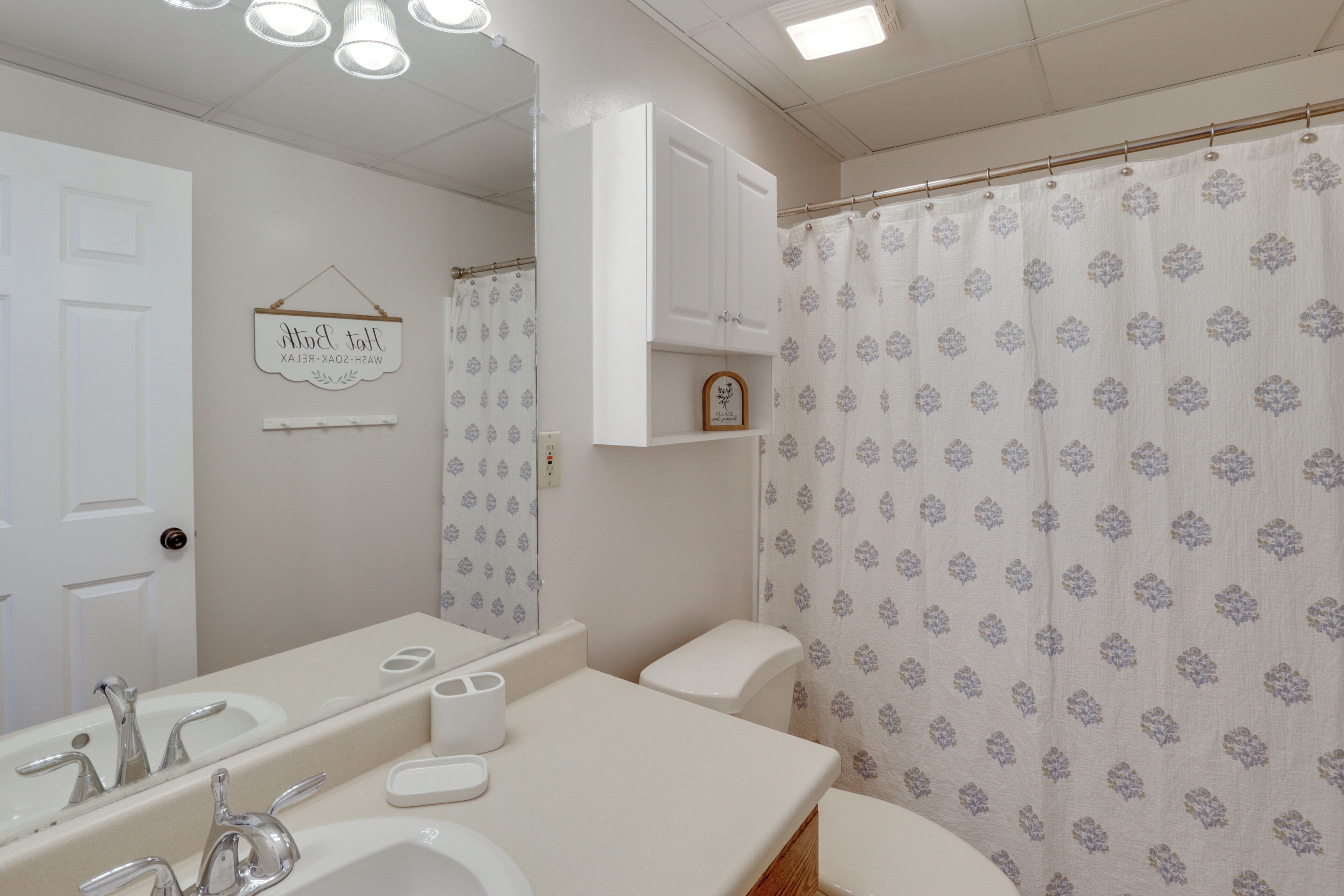 A bathroom with a white sink, mirror, and toilet. There is a shower curtain with a floral pattern and a sign above the mirror.