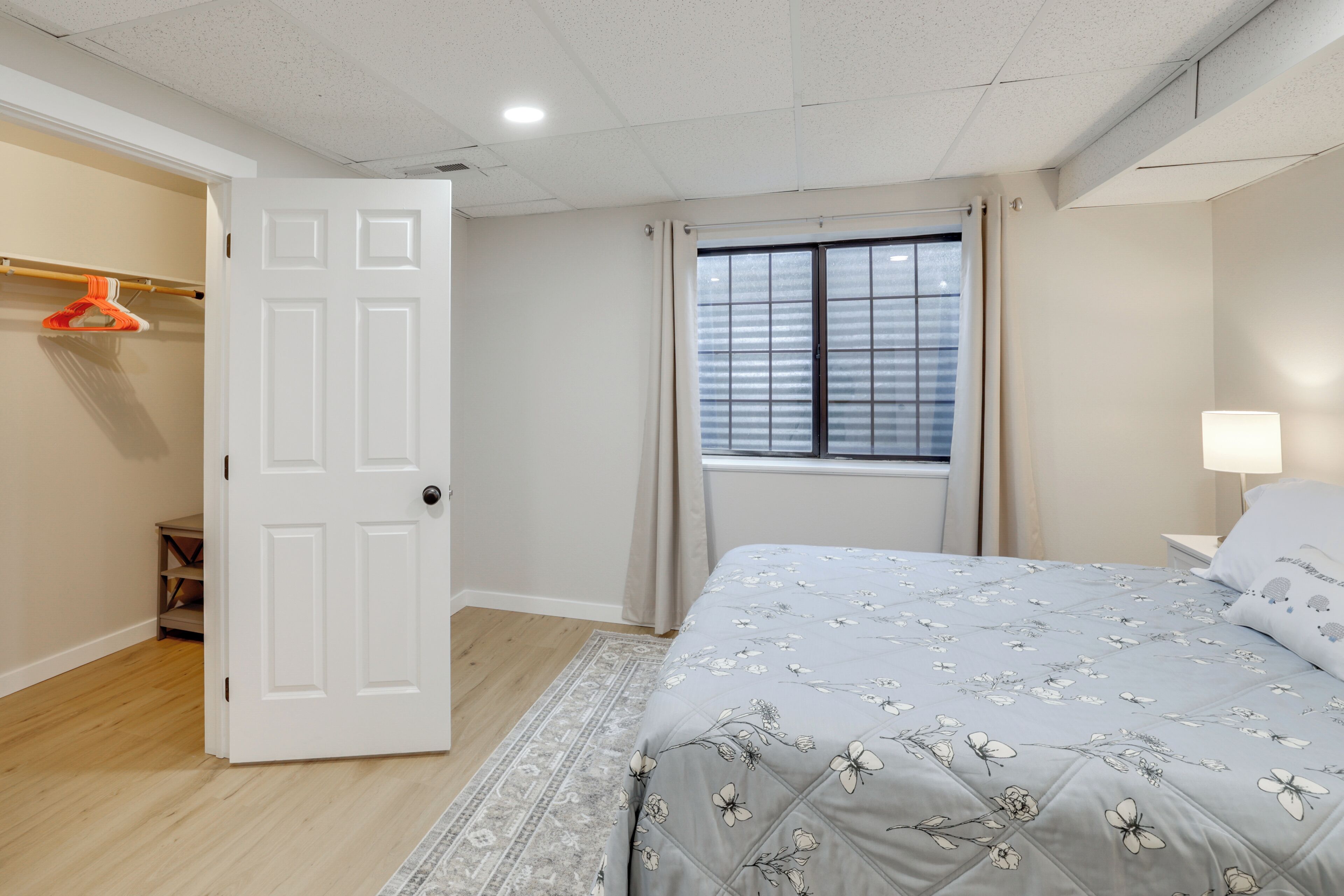 A bedroom with a bed, a window with blinds, a door, and a nightstand with a lamp.