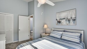 Un dormitorio con cama, ventilador de techo, mesita de noche con lámpara, un cuadro en la pared y una puerta que da a otra habitación.