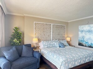 A hotel room with a bed, a blue sofa, a decorative wall panel, a potted plant, and a painting. - Rent Ready Beautiful Unit in Caravelle in Myrtle Beach! (Myrtle Beach)