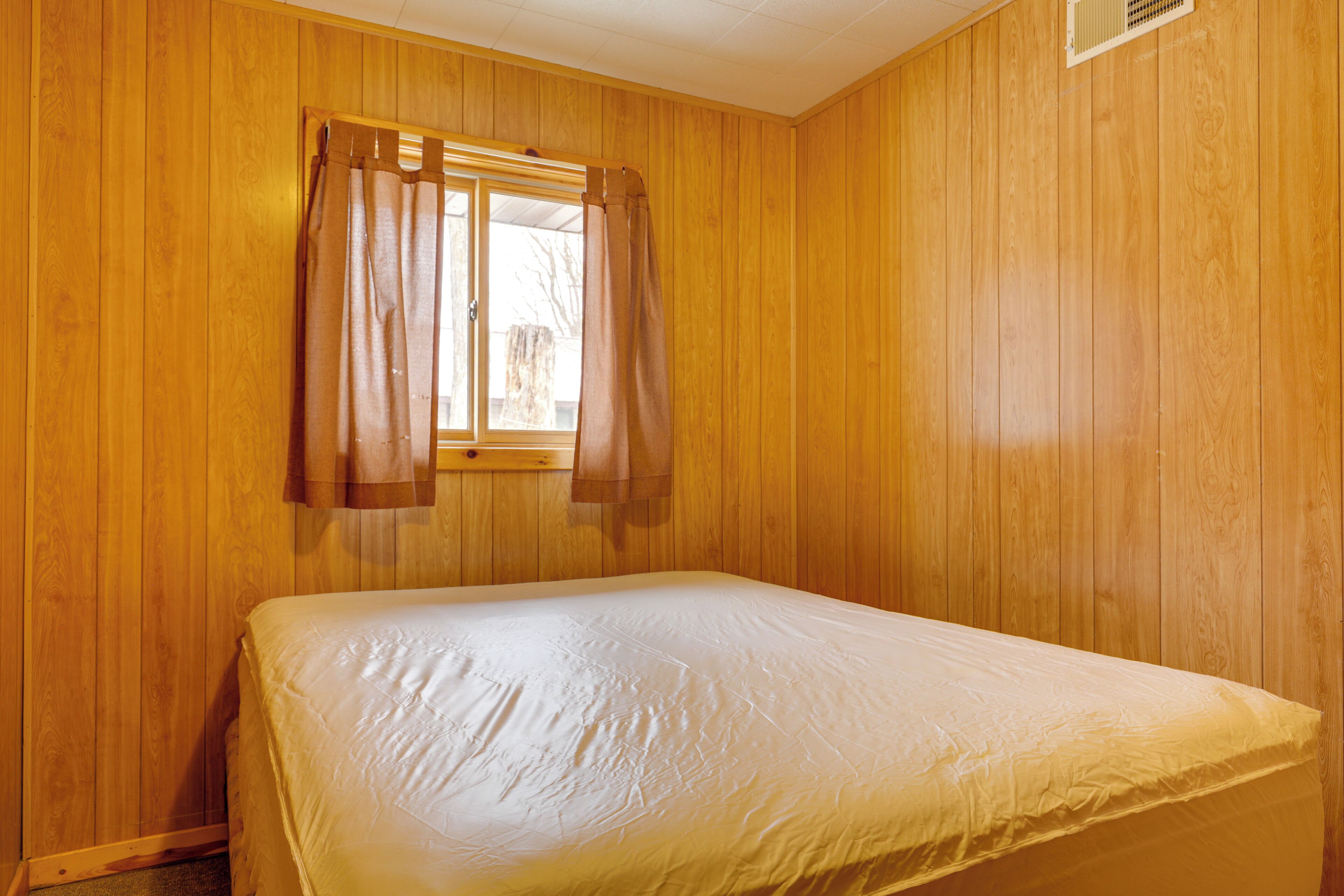 A room with wooden walls, a single bed with a white mattress, and a window with brown curtains.