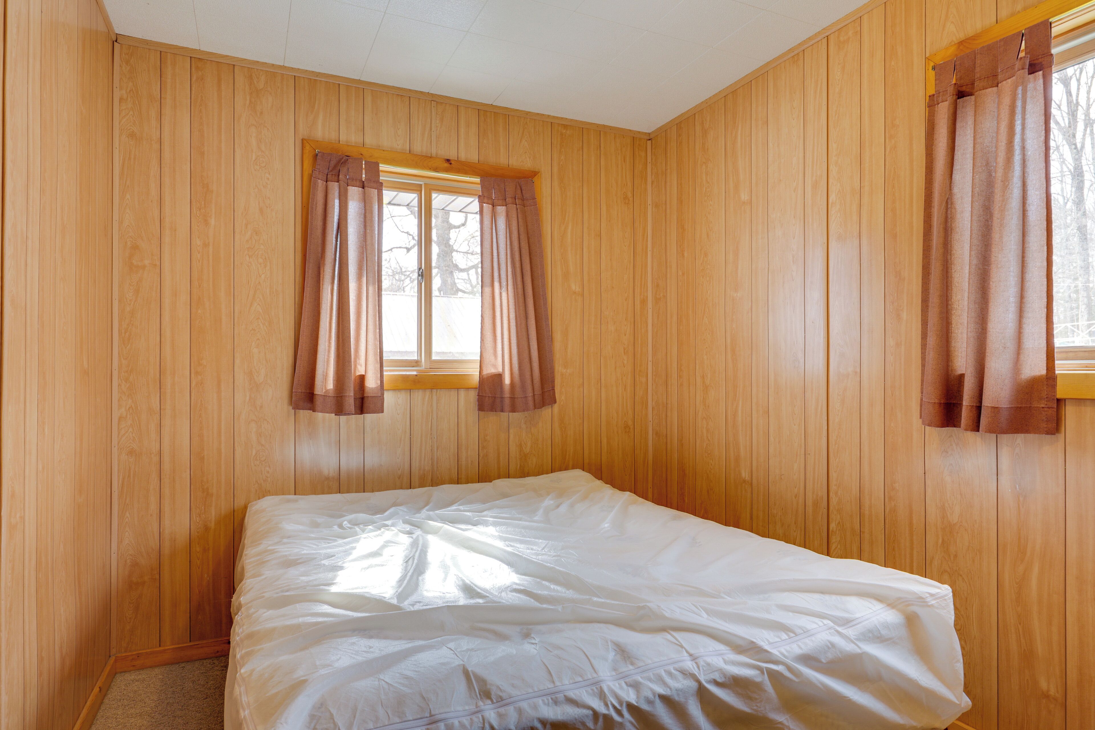 Ein Zimmer mit Holzwandverkleidung, einem Einzelbett mit weißer Bettwäsche und zwei Fenstern mit braunen Vorhängen.