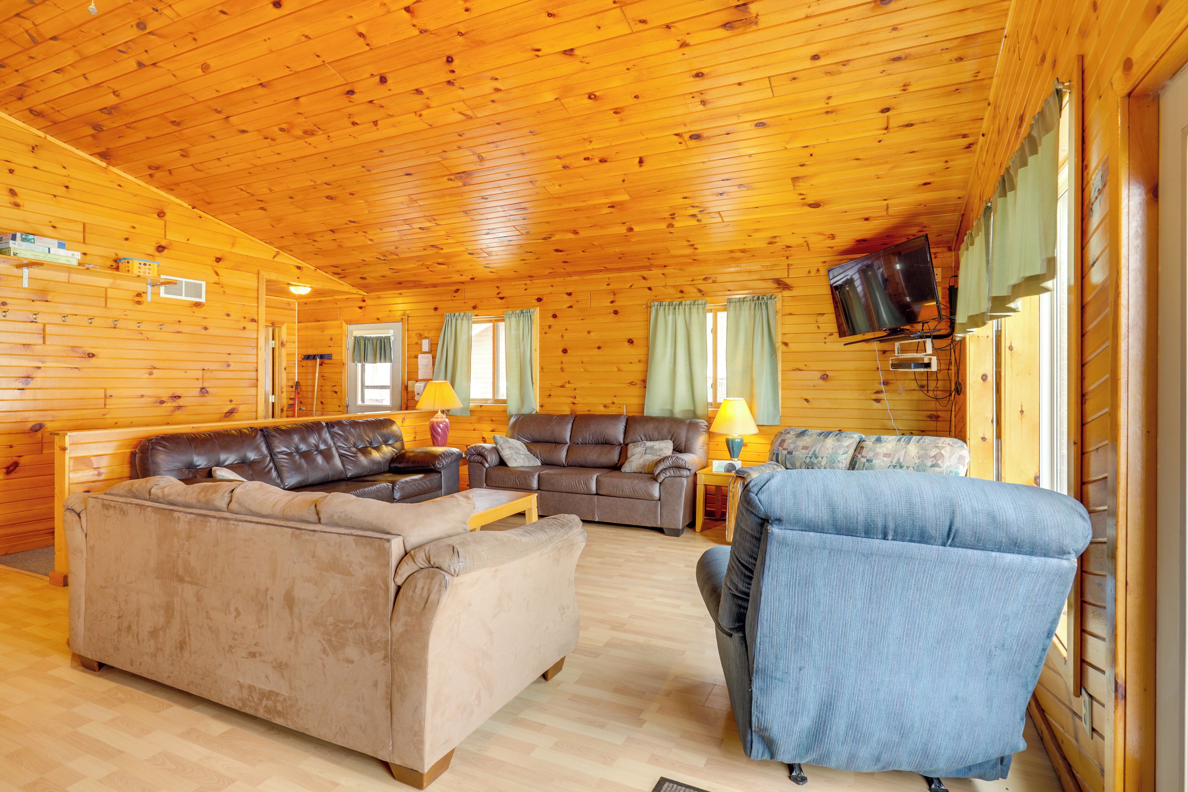 A log cabin interior with a wooden ceiling, a brown leather sofa, a blue armchair, and a television mounted on the wall.
