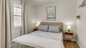 Une chambre à coucher comprenant un lit, deux tables de chevet, une commode, un ventilateur de plafond, une fenêtre avec des rideaux, un tableau accroché au mur et une petite étagère avec une plante.